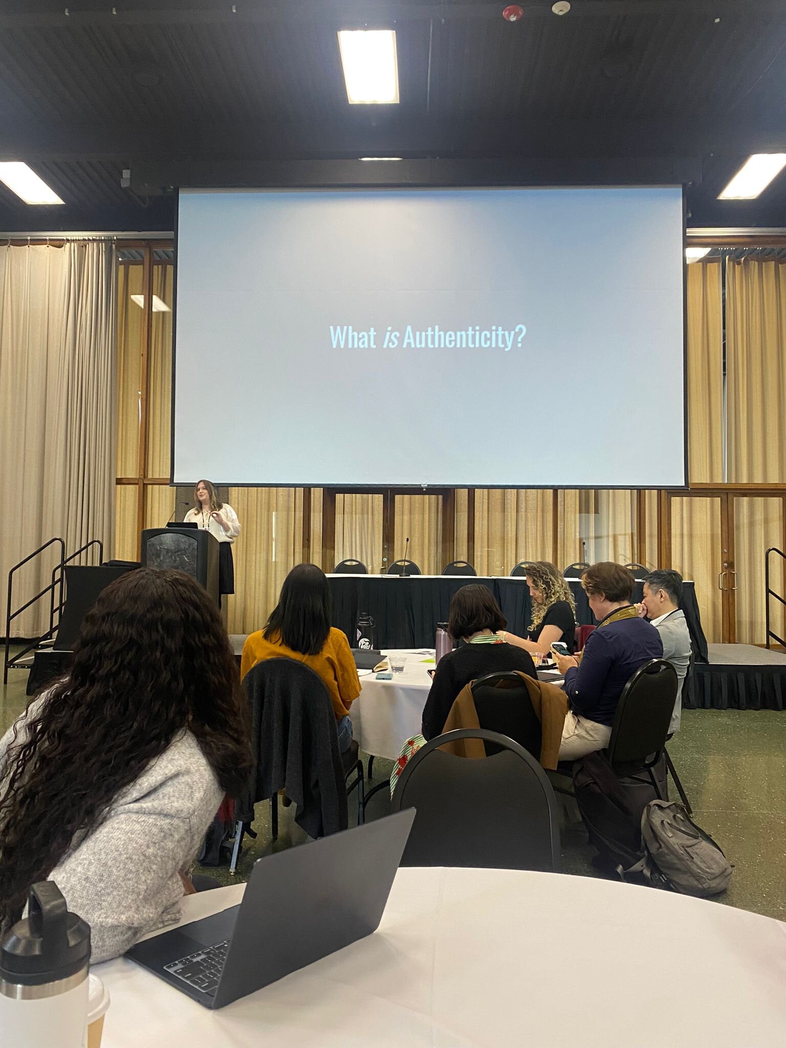 Logan McLaughlin presenting to a seated audience beside a slide reading "What is Authenticity?"
