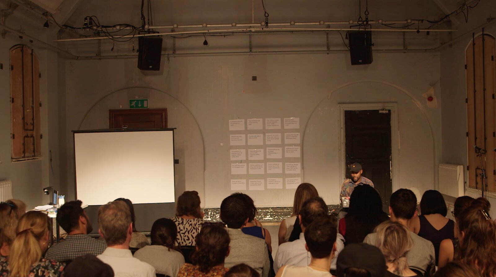 Harun Morrison addressing a seated audience beside a grid of question cards on the wall