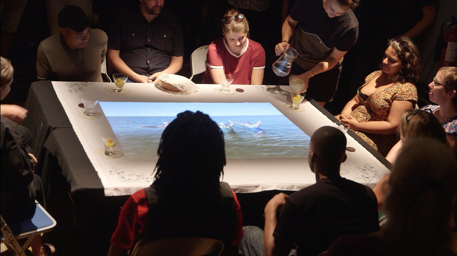 Audience gathered around a tablecloth-covered table with a projected seascape, in Rhiannon Armstrong's piece